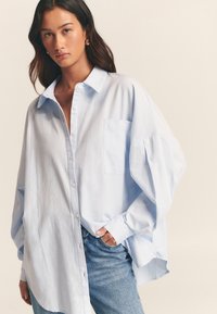 Light blue oversized button-up shirt made of soft fabric with a pocket on the left and rolled-up sleeves, paired with denim jeans.