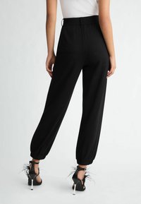 Pantaloni da jogger neri con una vita arricciata, caviglie con risvolto e dettagli goffrati sottili, abbinati a sandali neri con cinturini e dettagli in piuma.