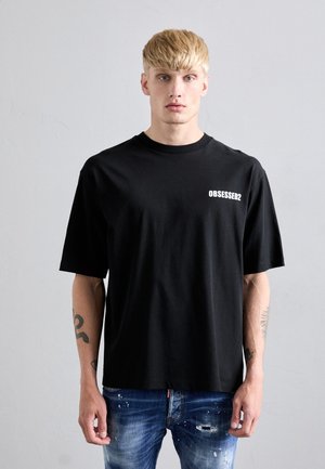 Uomo biondo con tatuaggi che indossa una maglietta nera con la scritta "OBSESSED2" e jeans blu strappati, in piedi contro una parete grigia uniforme.