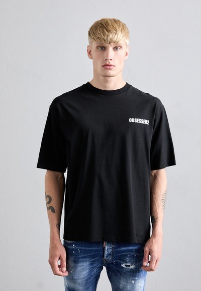 Blond man with tattoos wearing a black "OBSESSED2" T-shirt and distressed blue jeans standing against a plain gray wall.