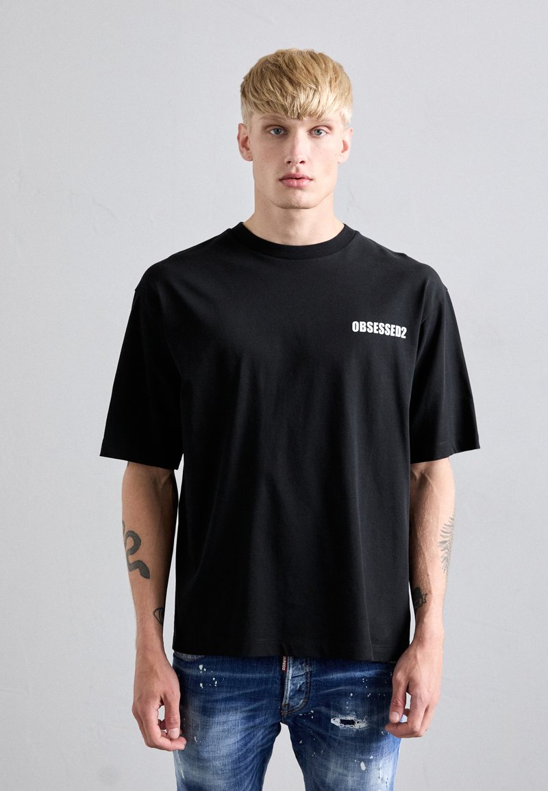 Uomo biondo con tatuaggi che indossa una maglietta nera con la scritta "OBSESSED2" e jeans blu strappati, in piedi contro una parete grigia uniforme.