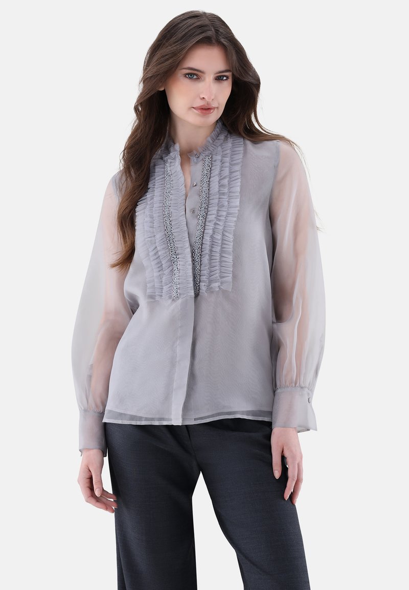Helle graue Bluse mit gerafftem Vorderteil, Knopfverschluss und transparenten langen Ärmeln. Der Stoff hat eine glatte Textur mit dezentem Glanz.