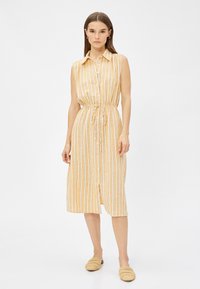 Vestido sin mangas de rayas amarillas con botonadura frontal y cintura con cordón. Fabricado en una tela ligera. Combinado con zapatos de tela beige.
