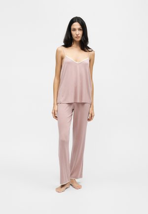 Femme portant un haut caraco en satin rose clair et un pantalon large assorti, debout pieds nus sur fond blanc.