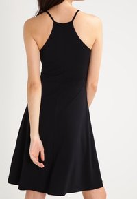 Donna che indossa un vestito nero senza maniche con spalline sottili, vista di spalle su uno sfondo chiaro e semplice.