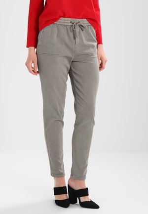 Woman wearing grey drawstring pants with front pockets, a red long-sleeve top, and black block-heeled mules standing against a white background.