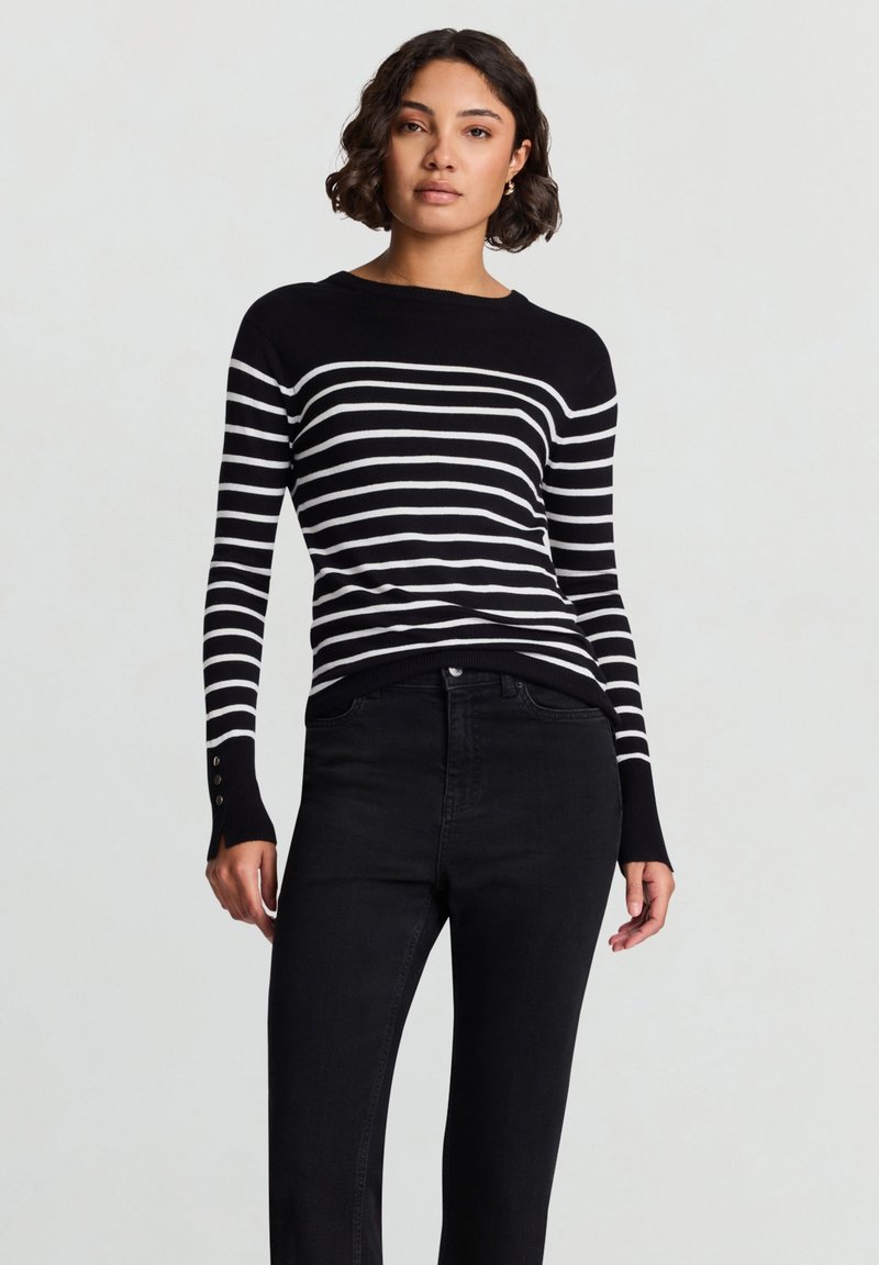 Schwarz-weiß gestreifter Strickpullover mit Rundhalsausschnitt, langen Ärmeln und Knopfverzierungen an den Bündchen, kombiniert mit schwarzer Jeans.