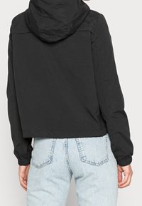 Černá zkrácená bunda s kapucí, elastickými manžetami a hladkým povrchem. Spárování s světle modrými džíny, které mají viditelné zadní kapsy.