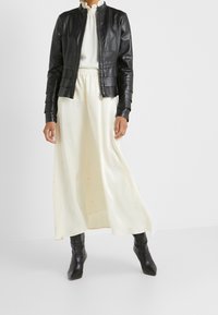 Black leather jacket with a fitted design, long sleeves, and pleated accents. Paired with a cream maxi dress featuring small black dots.