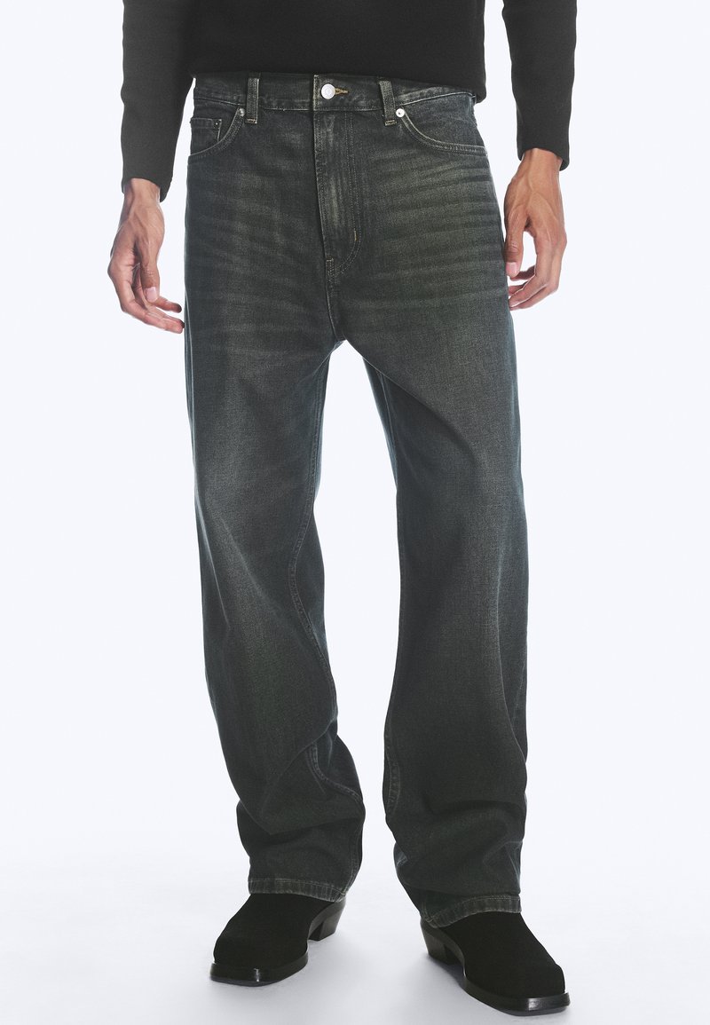 Homme portant une chemise noire à manches longues, un jean ample gris foncé avec un léger délavage et des bottes noires à bout carré, debout avec les mains détendues sur les côtés.