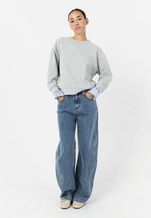 Nainen, jolla on tumma tukka ponnarilla, pukeutunut harmaaseen collegepaitaan siniraitaisen paidan päällä, väljät siniset farkut ja vaaleanbeiget kengät, seisoo käsivartensa taskuissa.