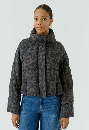 Frau mit einer dunkel gemusterten Steppjacke mit Leopardendruck und hohem Kragen sowie blauen Jeans, stehend vor einem einfarbigen hellen Hintergrund.