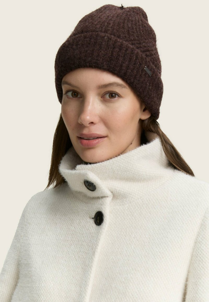 Bonnet en tricot marron avec un motif texturé, associé à un manteau blanc texturé avec un col haut et des accents de boutons noirs.
