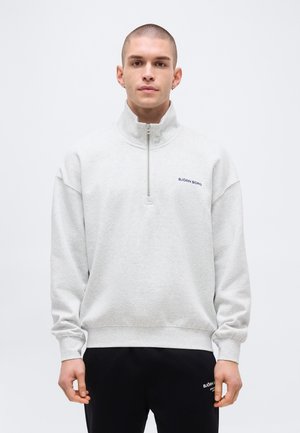 Jeune homme portant un sweat-shirt à quart de zip gris clair et un pantalon noir, tous deux avec des logos Björn Borg, debout devant un fond blanc uni.