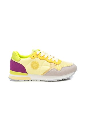 Zapatilla baja con paneles amarillos, beige y morados, cordones blancos y suela de goma, mostrada desde el lateral exterior sobre un fondo blanco.