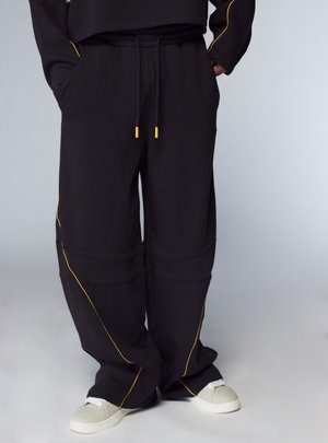 Pantaloni della tuta neri con dettagli in cuciture gialle, caratterizzati da una vestibilità ampia, vita elastica e tasche laterali; abbinati a scarpe da ginnastica bianche.