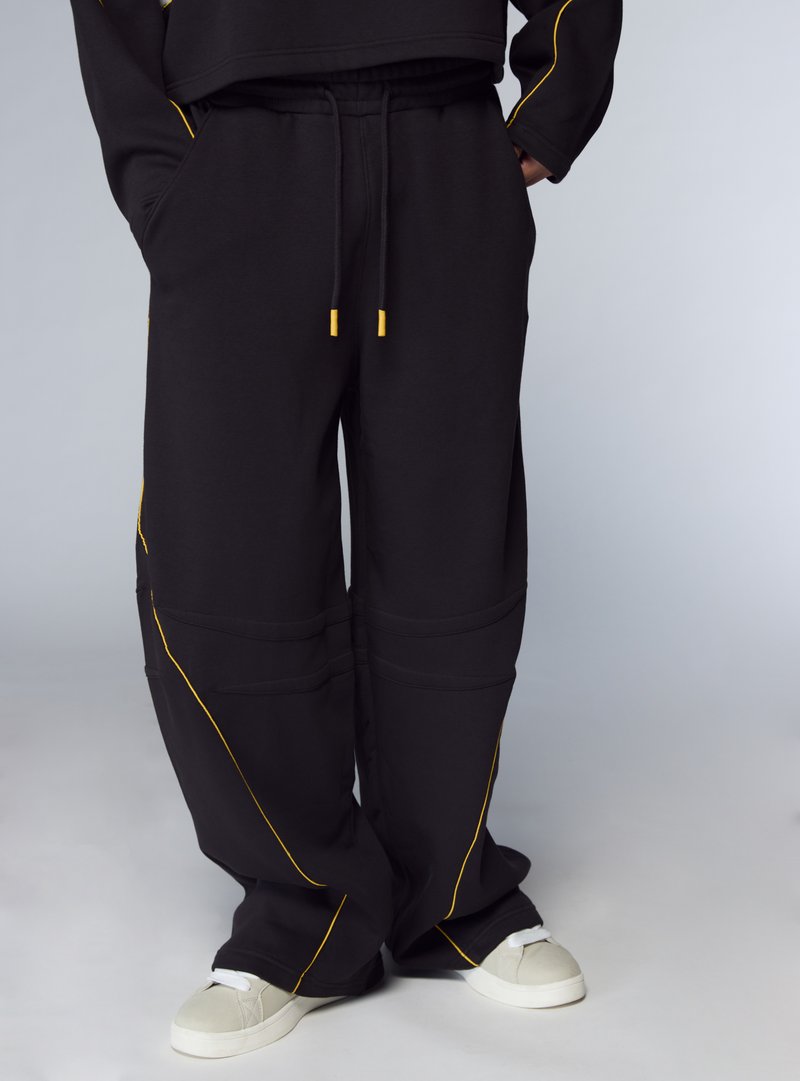 Pantaloni della tuta neri con dettagli in cuciture gialle, caratterizzati da una vestibilità ampia, vita elastica e tasche laterali; abbinati a scarpe da ginnastica bianche.