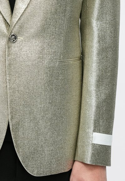 Metaliczna złota marynarka z fakturowanej tkaniny, zapinana na jeden guzik, z białym logo na mankiecie. Dopasowany krój i elegancki design.