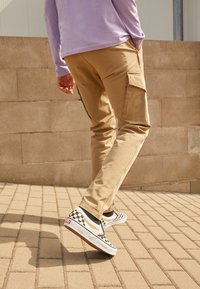 Beige cargobroek met zijzakken, gecombineerd met zwart-witte geruite slip-on schoenen, lopend op een geplaveide ondergrond, met een lichtpaarse lange mouwen top.
