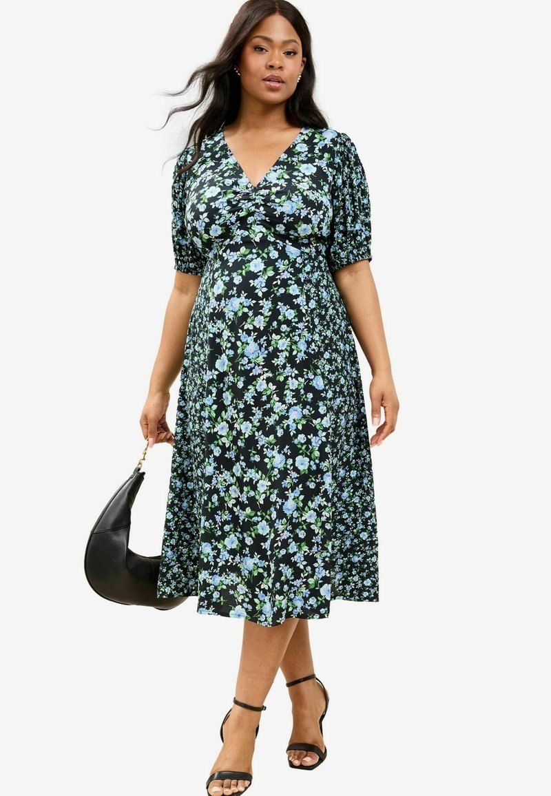 Schwarzes Blumenkleid mit kurzen Puffärmeln, verziert mit blauen und grünen Blumenmustern, und einer fließenden Silhouette. Kombiniert mit Riemchensandalen und einer schwarzen Tasche.