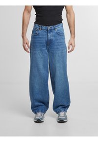 Jeans en denim bleu ample avec une coupe droite, cinq poches et une finition légèrement décolorée ; accompagnés de chaussures de sport grises.