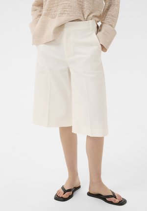 Osoba nosiaca voľné biele šortky po kolená, béžový textúrovaný dlhý rukáv a čierne šľapky so štvorcovou špičkou, ruky v vreckách.