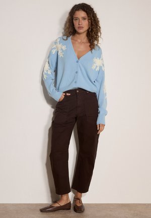 Frau mit lockigem Haar trägt einen blauen, geknöpften Pullover mit weißen Blumenmustern, dunkelbraune Hose und braune flache Schuhe, steht an einer Wand.