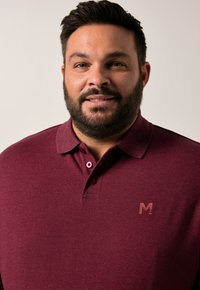 Muž s krátkými tmavými vlasy a plnovousem, oblečený v burgunské polokošili s malým logem na hrudi, mírně se usmívající na jednobarevném pozadí.