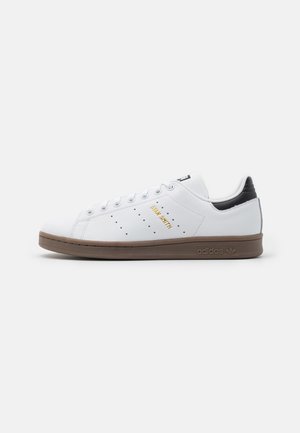Baskets blanches Adidas Stan Smith avec semelle marron, languette de talon noire, texte doré "Stan Smith" et trous de ventilation perforés sur le côté.