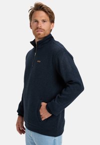 Navy fleece pullover with a half-zip, textured fabric, side pockets, and a leather label accent. Long sleeves with ribbed cuffs.