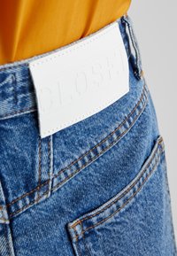 Detailní záběr na modré džínové kalhoty s viditelným oranžovým prošíváním a bílou koženou cedulkou na pase, v kombinaci se žlutým topem.
