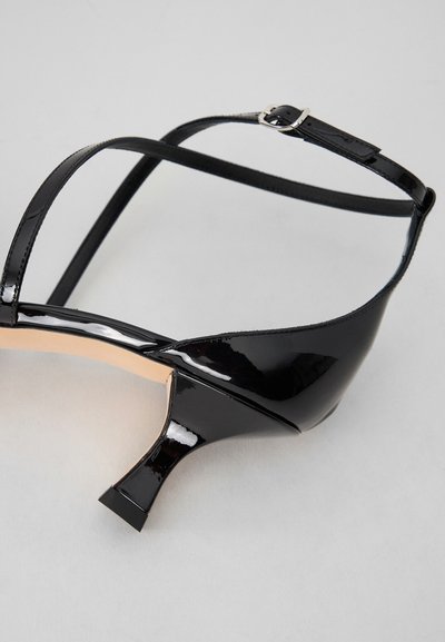 Sort laklæder hæl sandal med tynde stropper, spids tå og skulptureret firkantet hæl. Minimal hardware detalje med en ankelspænde.