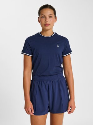 Jonge vrouw staat recht vooruit kijken, draagt een donkerblauw T-shirt met witte rand en bijpassende korte broek met elastische tailleband.