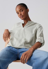 Personne assise portant une chemise à manches longues gris clair aux manches retroussées et un jean bleu, posant avec une posture détendue sur une chaise en bois clair.