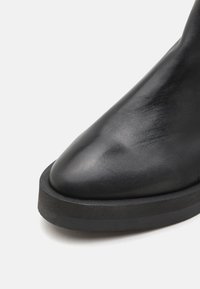 Bota de tobillo de cuero negro con punta afilada, que presenta una gruesa suela de goma negra y una textura suave.