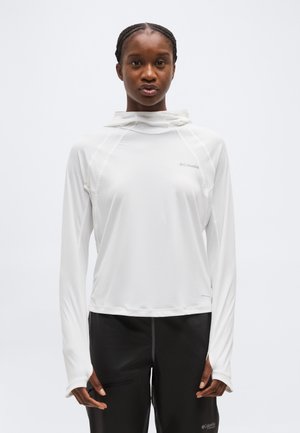 Άτομο που φοράει λευκό μακρυμάνικο μπλουζάκι με κουκούλα και τρύπες για τον αντίχειρα και μαύρο παντελόνι, μπροστά σε απλό ανοιχτόχρωμο φόντο.