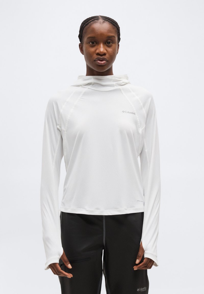 Άτομο που φοράει λευκό μακρυμάνικο μπλουζάκι με κουκούλα και τρύπες για τον αντίχειρα και μαύρο παντελόνι, μπροστά σε απλό ανοιχτόχρωμο φόντο.