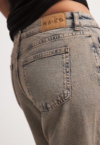 Gaišas džinsu auduma bikses ar teksturētu apdari, ar brūnu ādas uzlīmi ar uzrakstu "NA-KD" un redzamām šuvēm uz kabatām.