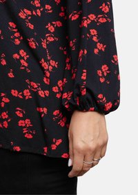 Schwarze Bluse mit floralem Muster, die rote Blumen zeigt, lange Puffärmel mit elastischen Bündchen und einem entspannten Schnitt. Nahaufnahme des Ärmels und der Handdetails.