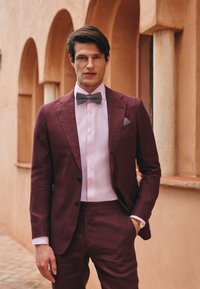 Completo bordeaux con vestibilità slim, abbinato a una camicia rosa chiaro e un papillon a fantasie. Notevoli dettagli del fazzoletto da taschino e della trama del tessuto.