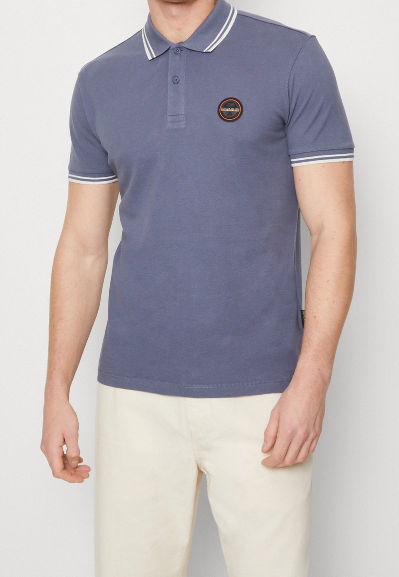 Polo-shirt bleu marin avec col et poignets rayés blancs ; muni d'une patte de boutonnage et d'un patch logo circulaire sur la poitrine. Matière en coton doux.
