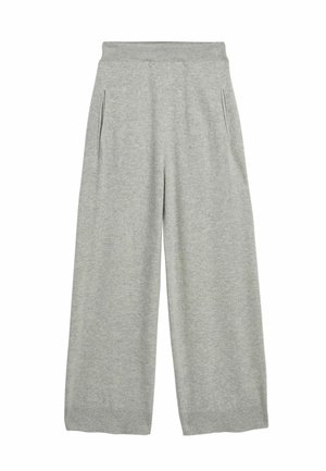 Tułów i biodra kobiety w jasnoszarej dzianinowej spódnicy i krótkim swetrze, ręka umieszczona w kieszeni spódnicy na neutralnym tle.