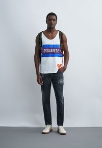 Homme portant un débardeur blanc à rayures bleues et violettes, un jean foncé, des baskets blanches et un sac à dos vert, debout devant un fond uni.