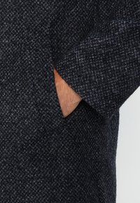 Abrigo de mezcla de lana que presenta un patrón de pied de poule gris oscuro, con un amplio collar y bolsillos laterales de tejido ligeramente texturizado.