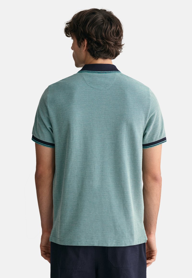 Mann steht mit dem Rücken zur Kamera und trägt ein helltürkises Polohemd mit dunklem Kragen und Ärmelborte, kombiniert mit dunklen Hosen.