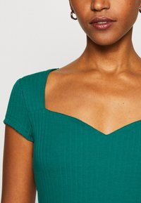 Teal geribbelde top met korte mouwen en een V-vormige halslijn. Gladde textuur met zichtbare stikdetails langs de naden.