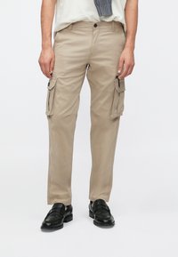 Μπεζ cargo παντελόνι με φερμουάρ στις πλαϊνές τσέπες, εφαρμοστή γραμμή, κούμπωμα με κουμπί και θηλιές για ζώνη· συνδυασμένο με μαύρα επίσημα παπούτσια.