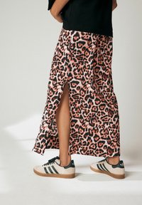 Jupe maxi en imprimé léopard rose avec une fente haute et un t-shirt noir, associée à des baskets beiges avec des accents et des rayures vertes.