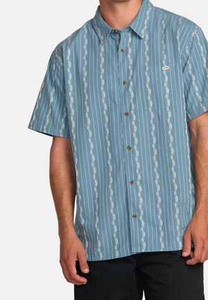 Camicia a maniche corte con bottoni di colore azzurro chiaro, ornata da strisce bianche ondulate e una piccola tasca sul petto. Presenta bottoni beige e una vestibilità comoda.