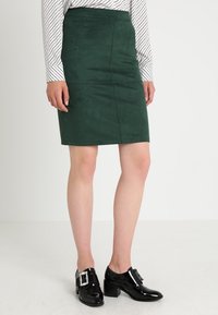 Grön mockapenskrid med vertikala sömmar; ihop med svarta lackskor med fyrkantig spänne.